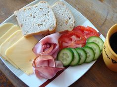 a white plate topped with bread, cucumbers and ham next to a cup of coffee