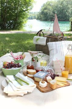 an outdoor picnic with cheese, fruit and juices on the table next to water