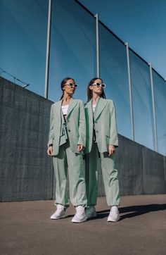 two women standing next to each other in front of a fence wearing green suits and white sneakers