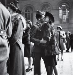 black and white photograph of two people kissing in front of a building with other people standing around