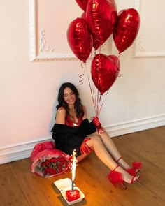 a woman sitting on the floor with red balloons and a birthday cake in front of her