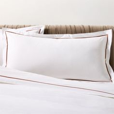 a bed with white sheets and brown piping on the pillowcase, in front of a beige upholstered headboard