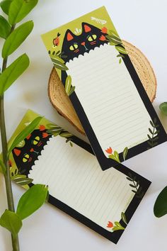 three notepads with black cats on them next to green leaves and a plant