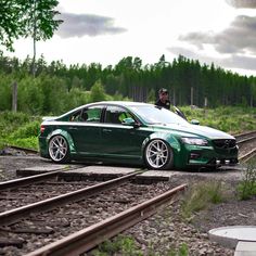 a green car parked on the side of train tracks next to a man in a black jacket