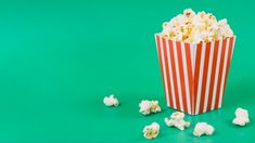 a red and white striped paper bag filled with popcorn on a green surface next to small pieces of popcorn