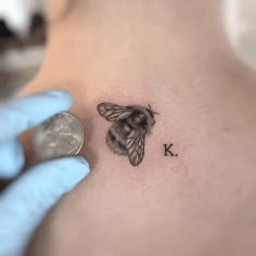 a bee tattoo on the back of a woman's left shoulder, with the letter k below it