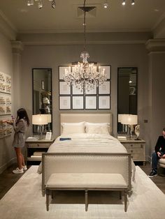 a woman standing in front of a large bed with a chandelier hanging from the ceiling