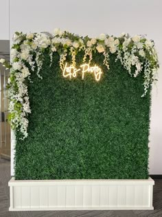 an artificial green wall with white flowers and greenery at the top that says let's party