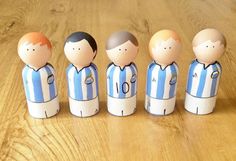 five small wooden dolls are lined up on a wood table, one is wearing a blue and white striped shirt