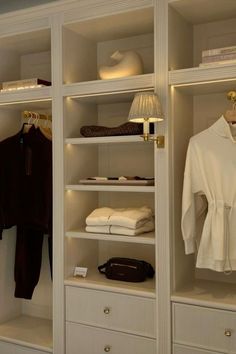 an open closet with white shelves and clothes on hangers, lamps and purses