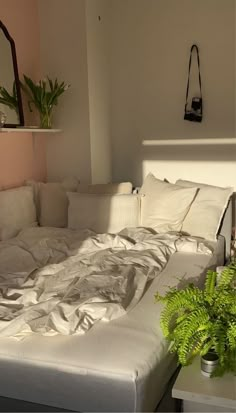 an unmade bed with white sheets and pillows in a living room next to a potted plant