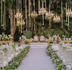 an outdoor wedding setup with white chairs and chandeliers hanging from the ceiling, surrounded by greenery