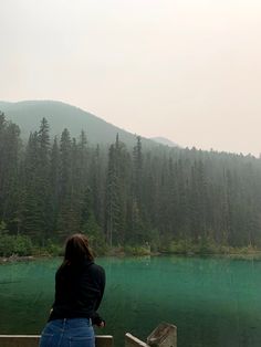 a woman sitting on a bench looking out over a lake in the woods with mountains in the background