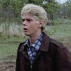 a young man with blonde hair wearing a brown jacket and blue checkered shirt is standing in the grass