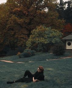 a woman sitting on the ground in front of a birdhouse and trees with orange leaves