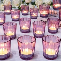 many purple glass cups with lit candles in them on a white tablecloth and flowers