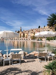 chairs and umbrellas are on the beach by the water with buildings in the background