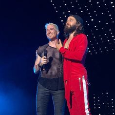 two men standing next to each other on stage with microphones in their hands and one wearing a red outfit