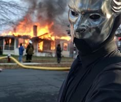 a man wearing a mask standing in front of a house on fire with flames behind him