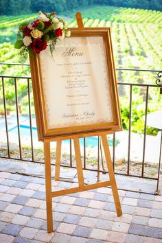 an easel with a menu and flowers on it sitting in front of a fence