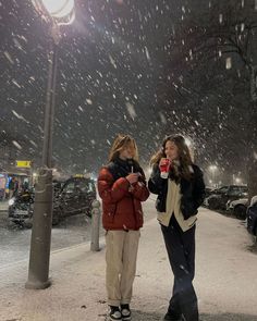 two women standing in the snow at night, one holding a coffee cup and talking