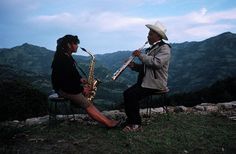 two people sitting on a bench with instruments in their hands and mountains in the background