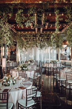 tables and chairs are set up for an event with greenery hanging from the ceiling