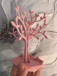 a hand holding a pink paper tree on top of a piece of art with flowers in the background