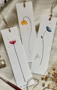 three bookmarks with watercolor flowers on them next to some scissors and yarns