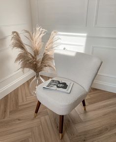 a white chair sitting on top of a wooden floor next to a vase filled with dry grass