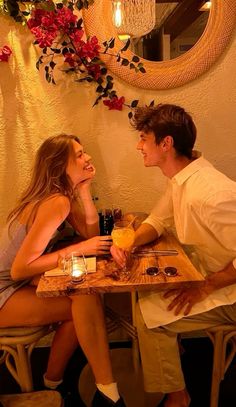 a man and woman sitting at a table with drinks in front of them, looking into each others eyes