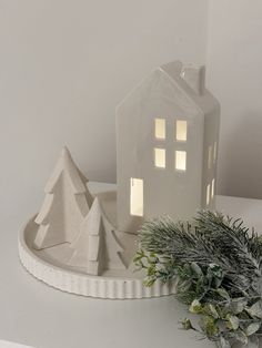 a small white house sitting next to a christmas tree on top of a table in front of a candle holder