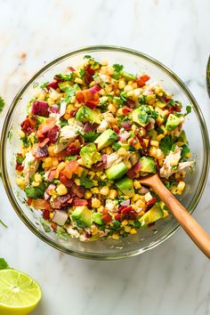 a glass bowl filled with corn salad and garnished with avocado, cilantro, red onion, and lime