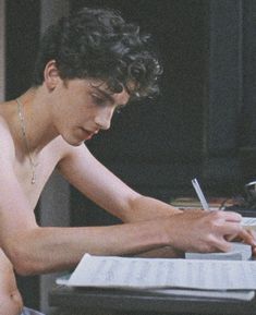 a shirtless young man sitting at a desk writing on a piece of paper with a pen in his hand