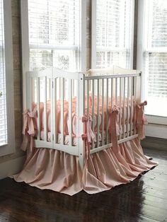 a white crib with pink ruffled bedding in front of two large windows