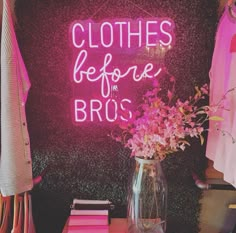 a pink neon sign that says clothes before the broc's next to a vase with flowers