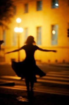 the silhouette of a woman dancing in front of a building at night with her arms outstretched