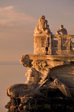 some statues are sitting on the edge of a cliff by the water and one is looking out to sea