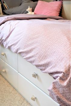 a white bed with pink and grey sheets on it's headboard, next to a book shelf