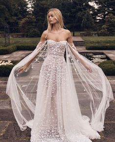 a woman wearing a white wedding dress with long sleeves and flowers on the skirt is standing outside