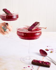 two glasses filled with red liquid and chocolate on top of a white table next to spoons