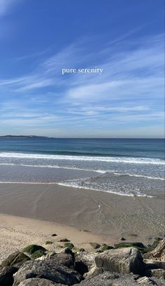 there is a rock on the beach with water in the back ground and blue sky above it