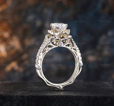 an intricately designed engagement ring sits on top of a black surface in front of a blurry background