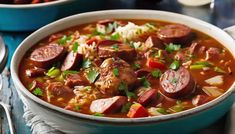 two bowls of soup with sausages, rice and carrots on the table next to silverware