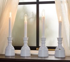 three white candles sitting on top of a table next to a window with curtains in the background