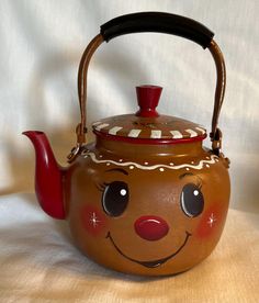 a brown tea pot with a red handle and face painted on the side, sitting on a white surface