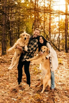 a man and woman are holding two dogs in the woods with leaves on the ground