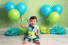 a baby sitting on the floor in front of some blue and green balloons while holding a toy