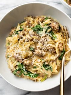 a white bowl filled with pasta covered in meat and spinach next to a gold fork