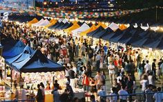 an outdoor market with lots of tents and people walking around it at night, in the evening